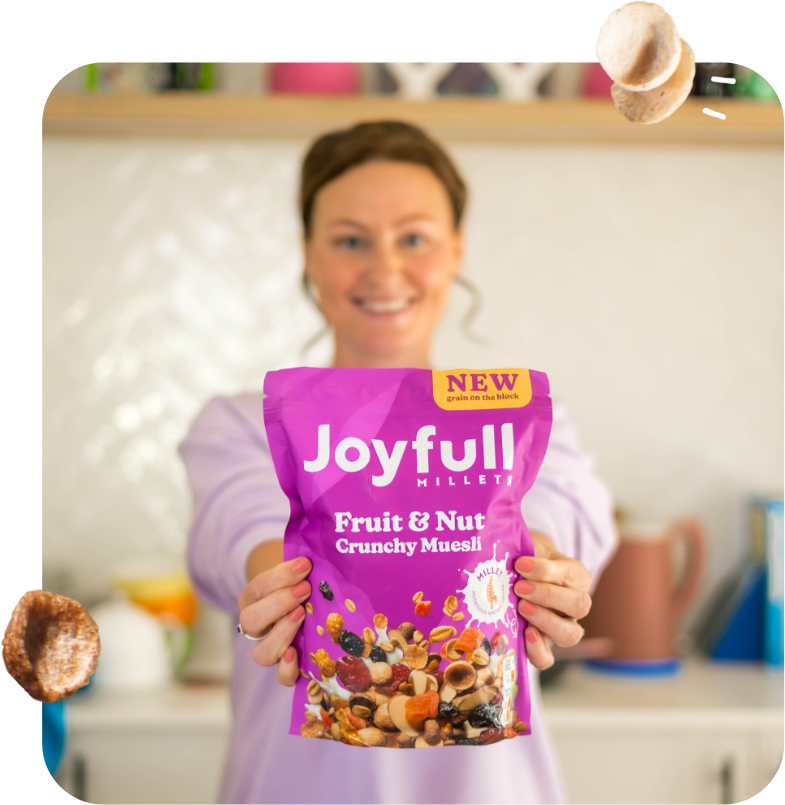 A picture of a woman holiding a pack of Joyfull Millets Fruit and nut cereal. The pack is purple, it has a splash of product in the bottom. On the top right corner, the pack has a yellow box that says "New". The woman in the background is out of focus, she is smiling and wearing a lilac sweater