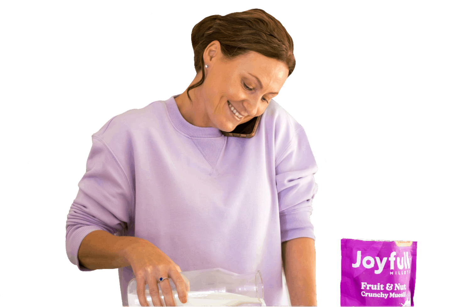 A picture of a brunette woman wearing a purpler crewneck sweater. She appears to be speaking on the phone, holding it between her cheek and shoulder. She is pouring milk with one hand and to her righ tthere is a pouch of Joyfull Fruit and Nut